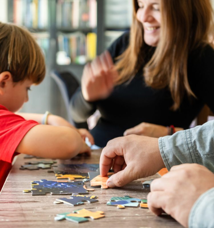 Focused boy assembling jigsaw puzzle with happy crop parents Side view of little boy playing wooden jigsaw puzzle at table with cheerful supportive parents at home